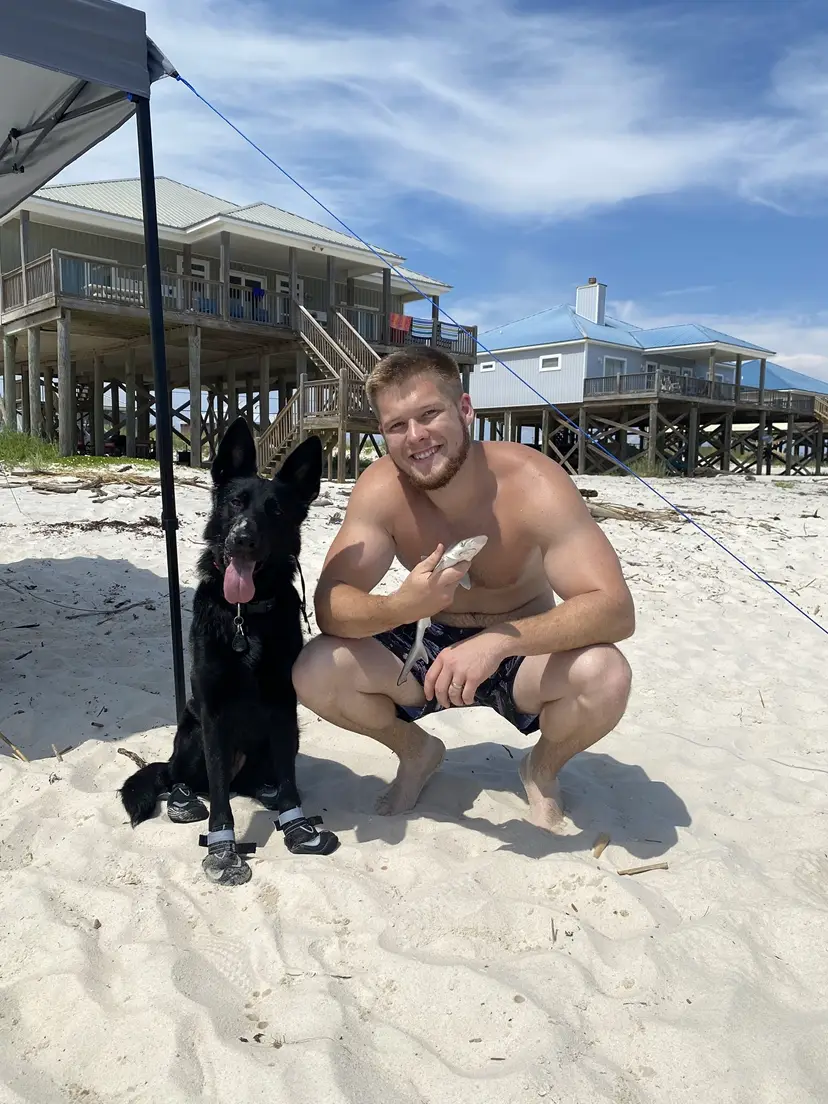 Bailey Clark crouching next to a black German Shepherd on a white sand beach