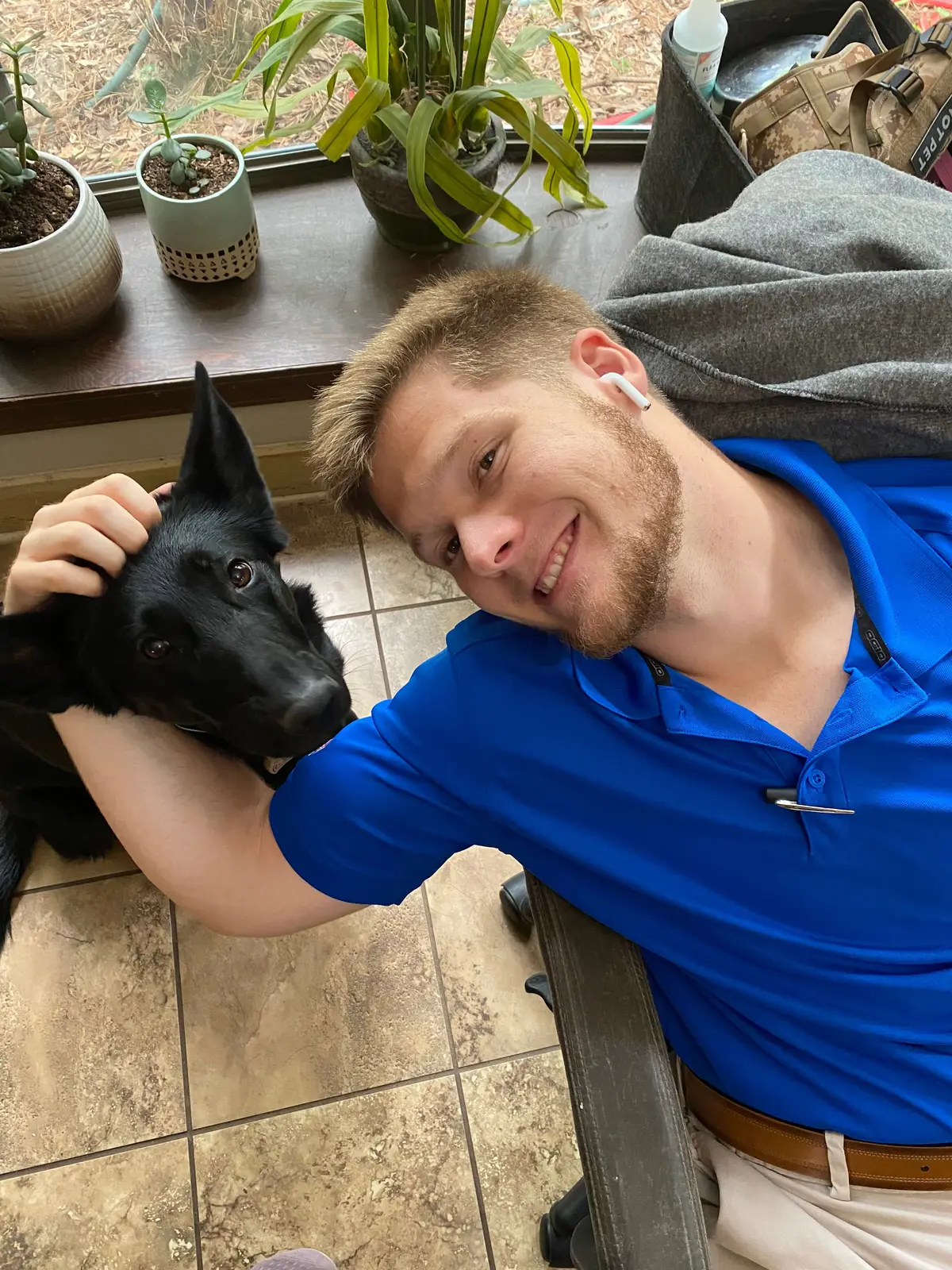 Bailey Clark smiling on the floor with a black German Shepherd resting its head on his arm
