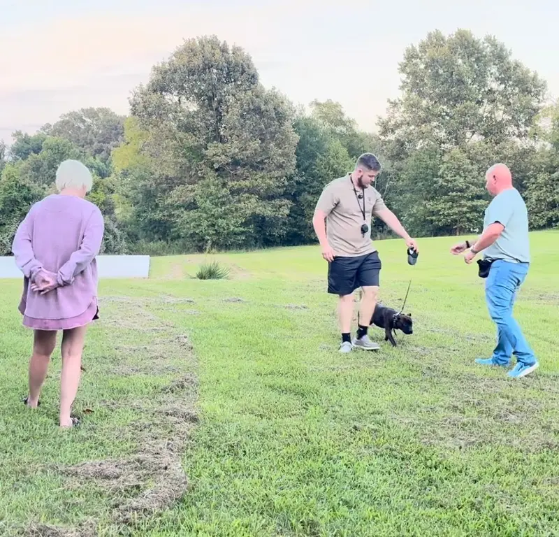 Bailey Clark working a small black dog on a long line in an open field while its owners watch during a private lesson