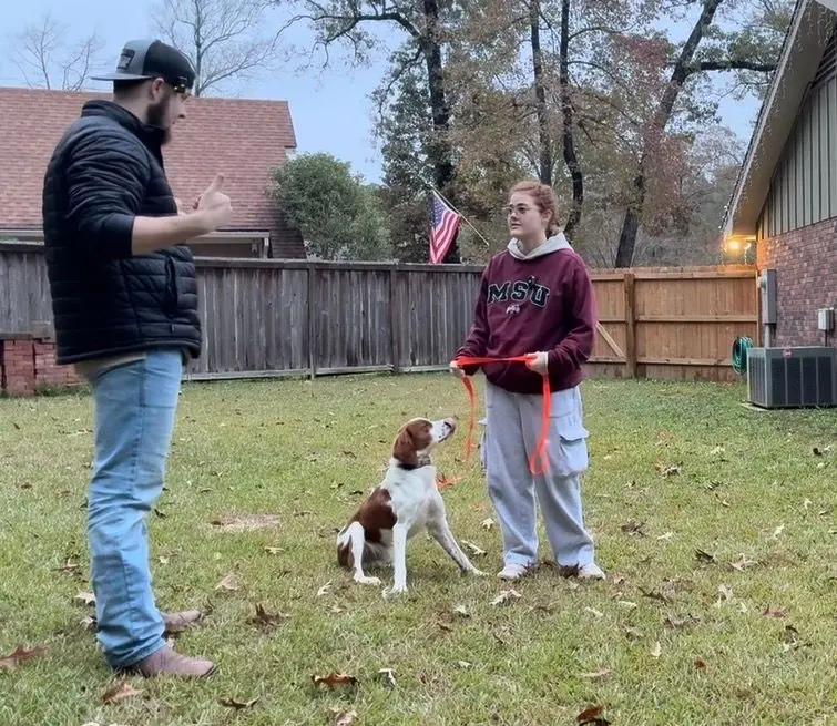 Bailey Clark coaching a young owner as her Brittany spaniel holds a sit on leash in their backyard