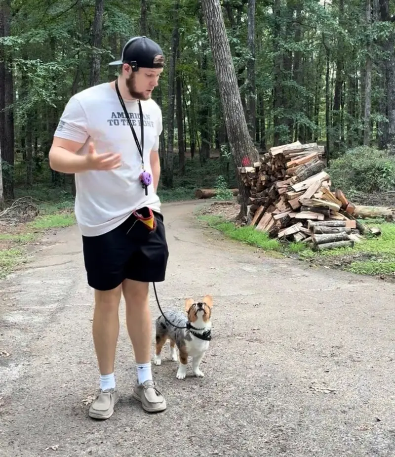 Bailey Clark coaching a corgi puppy on leash during a puppy training session on a wooded road